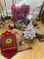Full view of white ceramic Christmas tree, plaid storage bag, assorted Christmas wreath and decorations on wooden floor