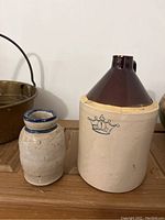 View of the two stoneware pieces side by side, showing the gallon jug crock with emblem and the smaller pottery jar.