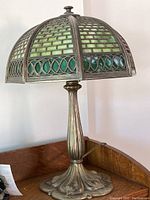 Full view of lamp on wooden surface showing cast metal floral base and stained glass dome shade with green and blue glass in metal framework.