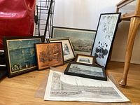 Image showing eight prints, framed and loose, leaning against the wall and on the floor. Various sizes visible, with different artwork styles including urban and botanical.