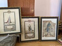Three framed prints leaning against a wooden door, showing two historical costume prints and one architectural drawing print, all framed in ornate gold frames.