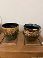 Two vintage Roseville pottery jardiniere planters, side by side on a wooden surface, showing brown and green glaze with floral designs