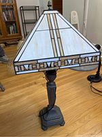 Front view of stained glass lamp showing square tapered shade with geometric stained glass design and decorative border, cast metal base with leaf motifs and hexagonal shape.