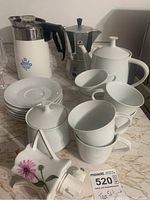 Photo showing cups and saucers stacked with teapot, sugar bowl with lid, silver espresso pot and floral container on marble countertop.