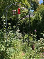 Photo of black metal garden trellis with two bird feeders hanging and a small hanging decoration at the base. Garden and green plants surrounding it in daylight.