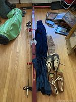 Photo of entire vintage ski set including two long wooden skis with bindings, bamboo poles, two pairs of leather ski boots, corduroy pants, gaiters, hat, and scarf laid out on wooden floor with other items visible in background.