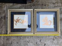 Two framed square art cards with native bronze art designs by Bill Reid laid on wooden surface with measuring tape showing about 13 inches size