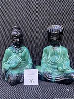 Two ceramic oriental figures with green and black glaze, one holding a fan and the other a book, photographed against a textured dark background.