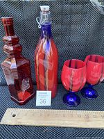 Photo showing two vintage glass decanters, one square red embossed and one tall red and blue swirl with stopper, alongside two red glass cups with blue stems, placed next to a ruler for size reference.