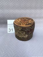 Photo of cylindrical wooden native box with bark sides and decorated slide top laid on textured background next to a MaxSold auction sign with lot number.