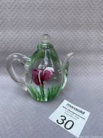 Clear blown glass teapot with green grass and purple-pink flower inset, shown front angle with handle right and spout left.
