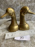 Pair of brass duck head bookends shown together from side angle against a textured backdrop with ruler for size reference.