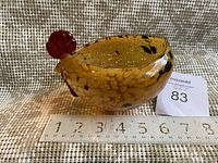 Blown glass chicken-shaped bowl viewed from the side with measuring tape showing length about 7 inches. Amber colored with dark spots, red comb and beak.