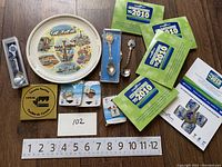 Overview of the entire lot showing several collectible items including the large world fair plate, collector spoons in boxes, medallion, pins, and Olympic pamphlets.