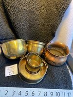 Five vintage metal bowls and pots arranged on black fabric with a ruler for size. Showing variation in size and metal types.