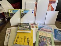 Photo showing multiple new white stretched canvases in original packaging of different sizes, several preowned artist and coloring books stacked in front.