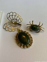Three vintage gold plated pieces against white background: crab pin with green jade stone, butterfly pin with filigree wings, and large oval jade pendant with ornate gold plated border.