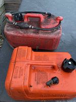 Two boat gas tanks side by side; the larger red tank shows attached fuel line and primer bulb hose, while the smaller orange tank lacks a fuel line.