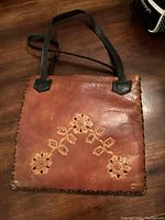 Slim rustic rust brown tooled leather purse with floral design on front and black shoulder handles shown on wooden surface.