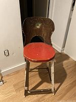 Front view of child's metal chair showing red painted circular seat with significant paint chipping and wear. Brown backrest with peeling paint and visible cracks.