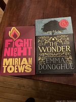 Two hardcover contemporary fiction books, Fight Night by Miriam Toews and The Wonder by Emma Donoghue, displayed on a wooden table.