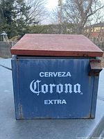 Front side of cooler with text 'Cerveza Corona Extra' on blue metal body and reddish wooden lid.