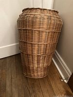Photo of the wicker basket standing on wooden floor beside white wall, showing its tall, narrow vase-like shape and natural brown weave