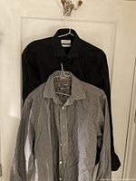 Pair of two men's dress shirts hanging on hangers on a white door. One is solid black, the other black and white striped. Shirts shown full length front view.