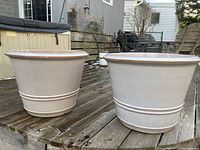 Two large beige ceramic plant pots placed on a wooden deck outdoors. Both have a smooth beige glazed surface with horizontal ring detailing near the base.