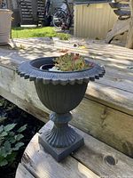 Single vintage cast iron plant pot on wood deck with ornamental scalloped rim and decorative fluted body, containing a black plastic liner and plant (plants not included).