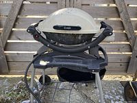 Closed Weber Q100 barbecue showing white lid with black handle, black base with stand and wheels outdoors on stone surface against wooden fence.