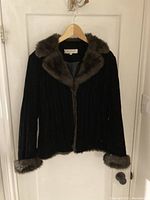 Front view of black women's jacket with brown faux fur collar and cuffs on a hanger against a white door.