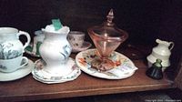 Shelf with various china cups, saucers, pitchers, and a colorful plate with a pink lidded dish.