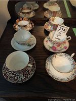 Six teacups with matching saucers arranged on a dark wooden surface, showing a variety of colors and floral decor.