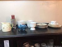 Wide view of items arranged on top of cabinet including pitcher, candleholder, fish lighter, milk glass bowls and plates