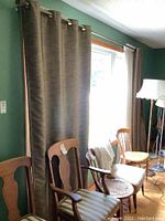 Two brown curtain panels hanging on a window with green walls, with visible wooden and cane chairs in front.