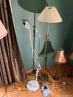 Photo of a 58-inch gold-colour metal floor lamp with wood trim and pleated cream shade next to a white metal and chrome adjustable arm floor lamp, with a 24-inch corded wall lamp visible nearby.