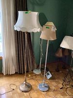 Two floor lamps standing side by side near a curtain. Left lamp is brass with white scalloped fabric shade, right lamp is metal with beige shade.
