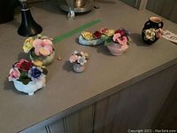 Photo showing six floral porcelain decor pieces arranged on table. Includes a variety of base shapes and colors, revealing detailed porcelain flowers.