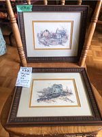 Two framed watercolour prints by Robert James Callaghan shown on wooden chairs. One print depicts a rural farm scene, the other a maritime scene, each in dark wooden frames with matting.