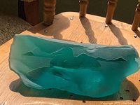 Photo showing the blue-green glass bird sculpture from the side and top angles on a wooden chair, revealing its shape and texture.