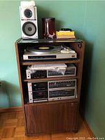 Full view of Fisher Studio Standard stereo equipment stacked inside wooden media cabinet with glass top. Turntable on top shelf, other stereo units below, AKAI speakers on top and side.