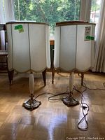Two tall brass, glass, and marble table lamps with white scalloped fabric shades standing on a wooden floor in front of a window.