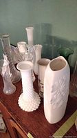 View of assorted vases on wood surface including milk glass, clear, and green glass pieces.