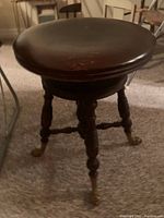 Full view of antique wooden piano stool showing round seat, spooled legs, claw feet with brass caps, and connecting stretchers.