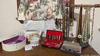 Wide photo showing assorted necklaces hanging on a wooden stand, jewelry storage boxes, porcelain teapot, and decorative floral basket on a wooden surface.