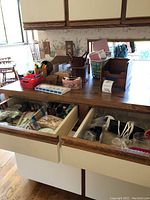 General view of kitchen drawers and countertop showing various kitchen utensils, wooden items, containers, and stationery supplies in baskets and boxes.
