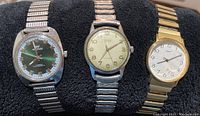 Three men's wristwatches displayed on black cushion showing Waltham (green dial), Gruen (off-white dial), and Timex (white dial) watches with metal bands.