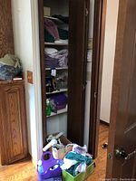 View of a closet with wooden shelves containing folded towels, rags, miscellaneous bottles, and a box at the bottom holding hair dryers and other items.