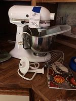 Front and partial side view of the KitchenAid Ultra Power mixer with mixing bowl attached, attachments and recipe booklet on wooden surface